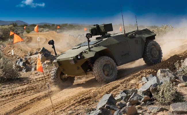 Armored all-terrain military vehicle driving over rocky desert terrain with orange flag markers.
