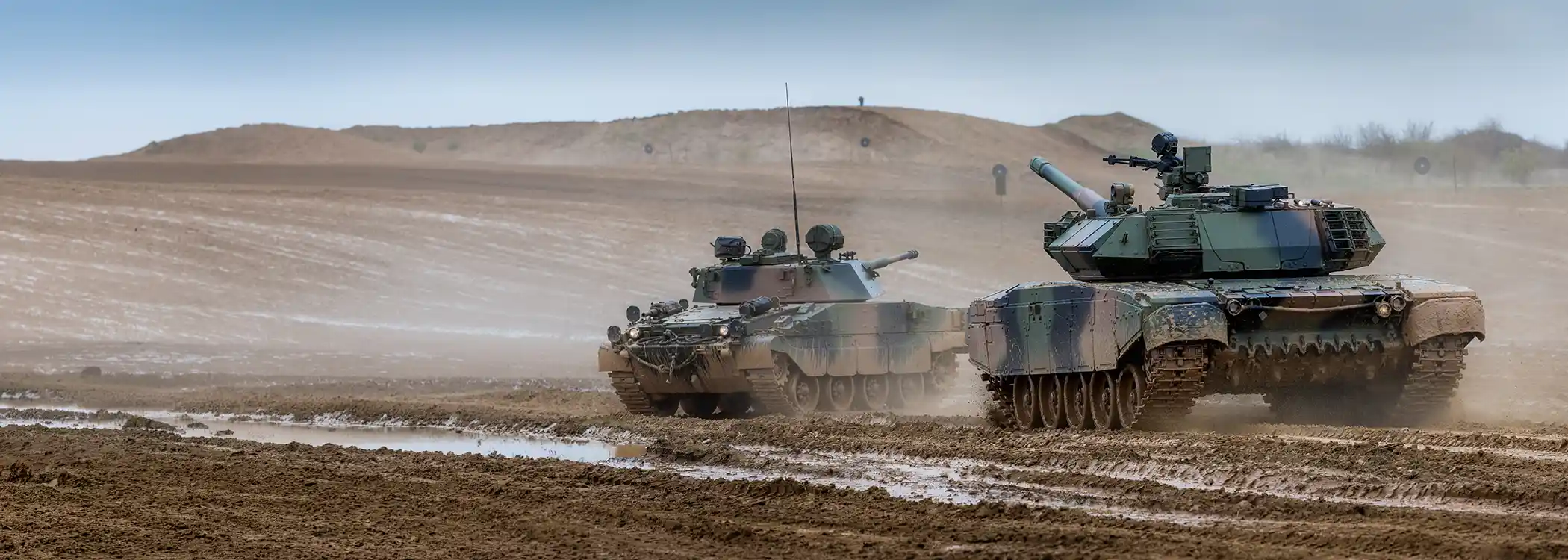 Two armored battle tanks rumble across a muddy desert landscape, kicking up dust as they advance in formation.