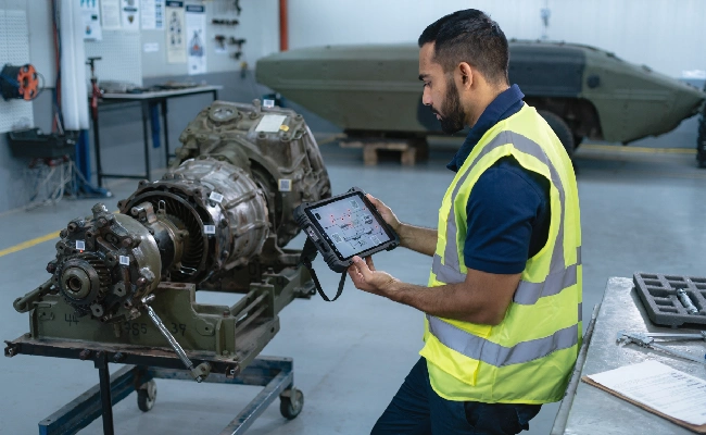 Male technician in a high-visibility vest inspecting a large gearbox on a wheeled stand using a tablet in a maintenance hangar with an aircraft fuselage visible in the background.