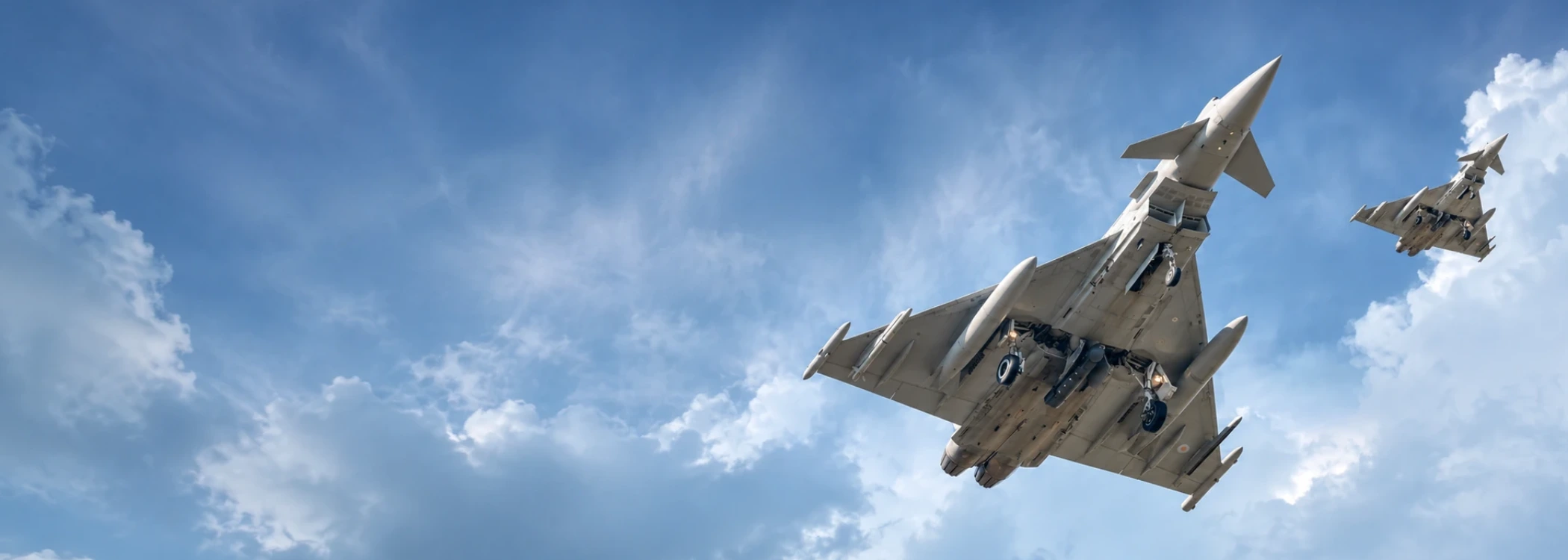 Two gray fighter jets fly in formation through a bright blue sky with clouds.