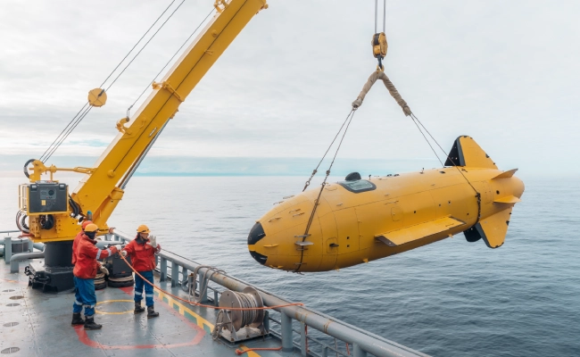 Yellow submarine-like submersible being hoisted by a crane from a ship deck, two workers in orange helmets guiding it toward the water.