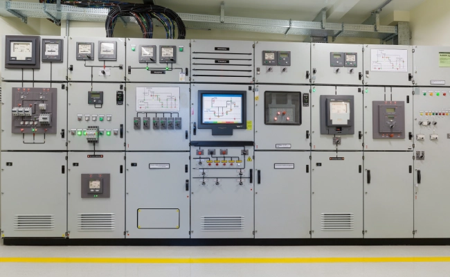 Row of industrial control panels and screens in a utility room, with wiring on top and yellow floor line nearby.