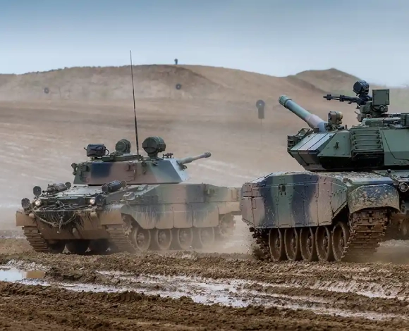 Two armored tanks advance through a muddy desert, dust rising from their tracks as barren hills loom in the distance.