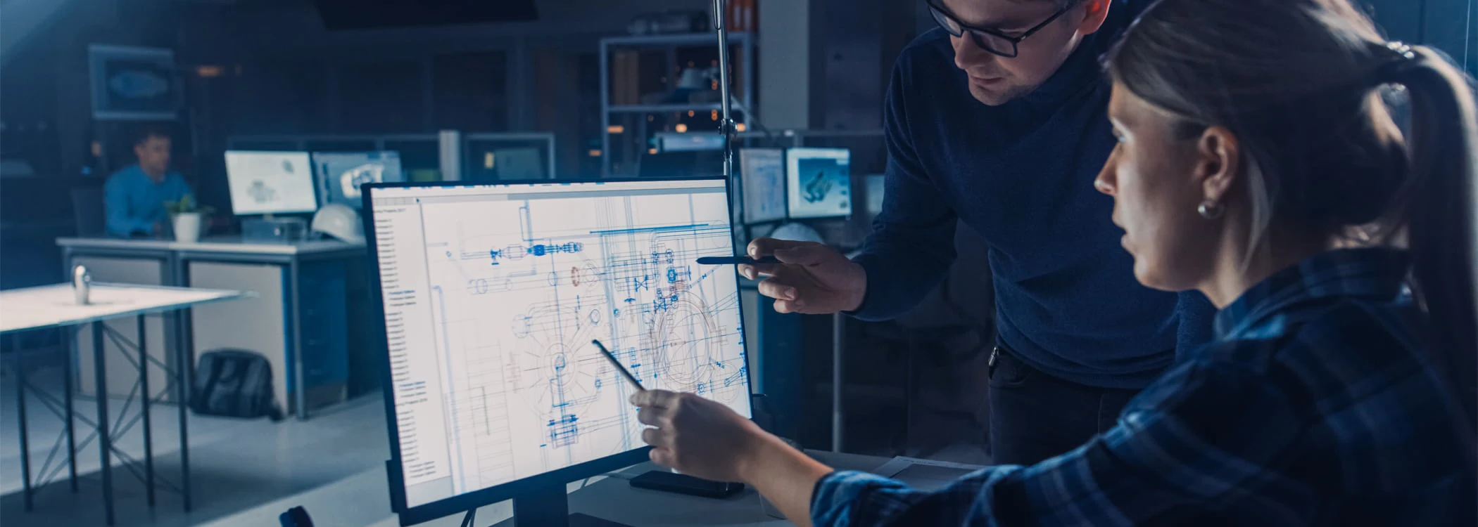 Two engineers review a detailed blueprint on a computer monitor in a dim, blue-tinted engineering lab.