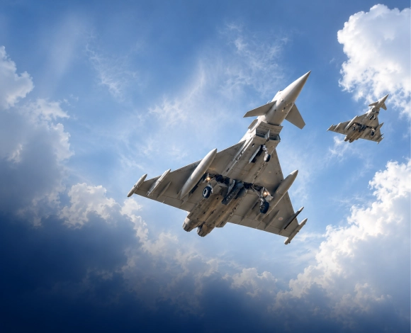 Two military fighter jets fly in formation against a blue sky with clouds, viewed from below.