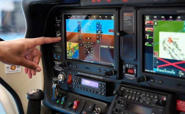 Pilot's hand points at a glass cockpit display showing attitude indicator and navigation map inside a small aircraft cockpit