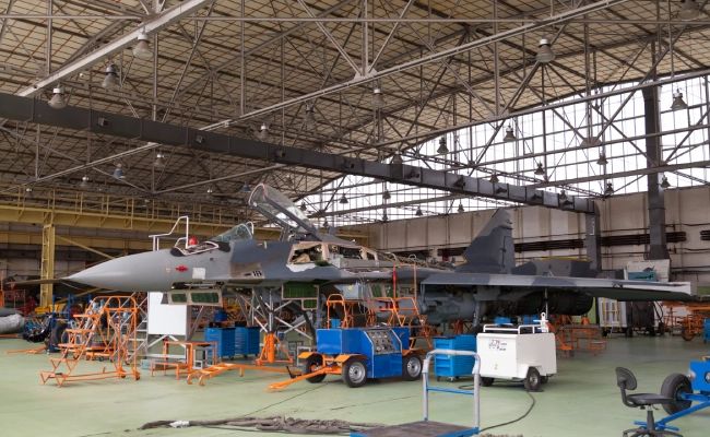 Military fighter jet undergoing maintenance inside a large hangar, surrounded by orange maintenance platforms and tools behind scaffolding.