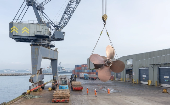 Port scene with a large crane hoisting a bronze ship propeller over a dock, workers in orange vests nearby.