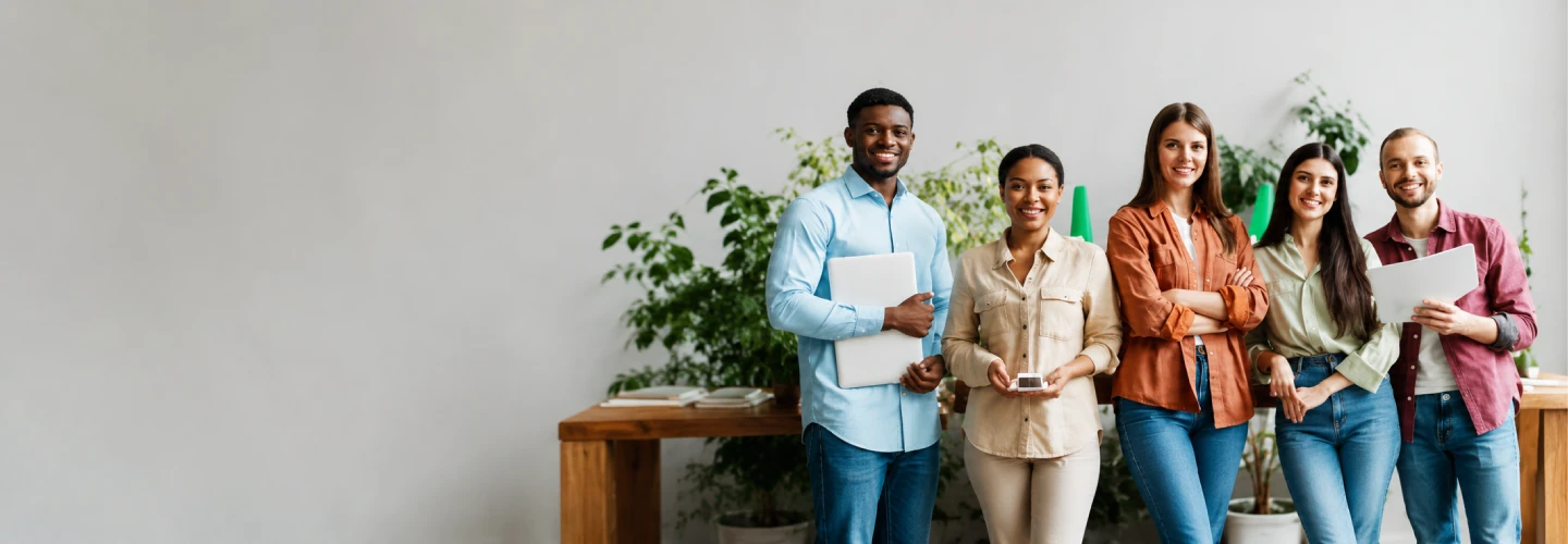 Five diverse coworkers stand together in a bright office, smiling, holding documents and a phone.