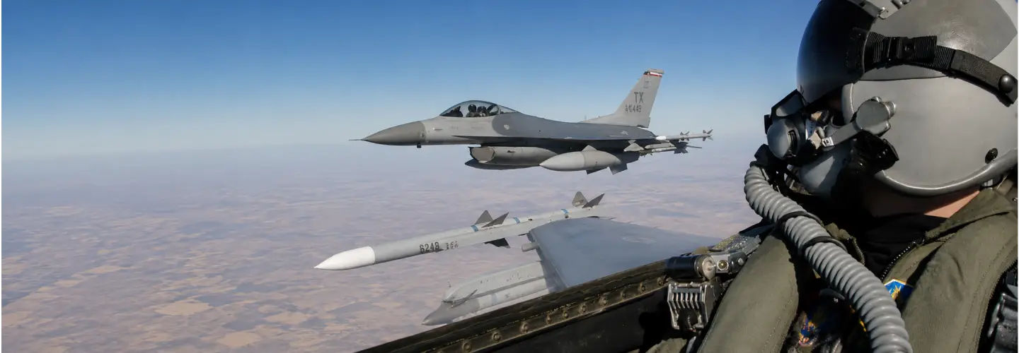 In-cockpit view of a fighter jet with another F-16 flying nearby and a missile under the wing, over a desert landscape.