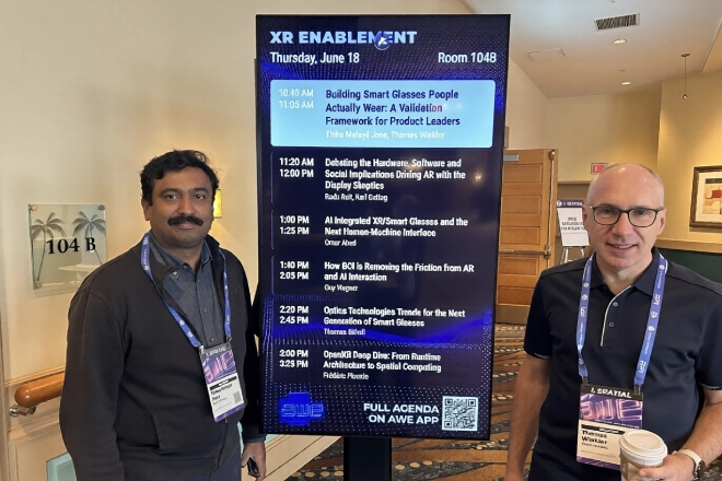 Two men with conference lanyards stand beside a tall digital schedule board titled 'XR ENABLEMENT' listing session times and topics.