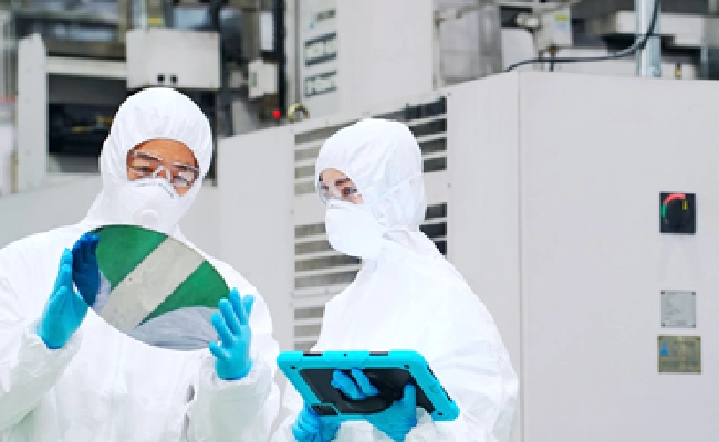 Two scientists in cleanroom suits inspect a circular silicon wafer, one holding it while the other uses a teal tablet nearby.