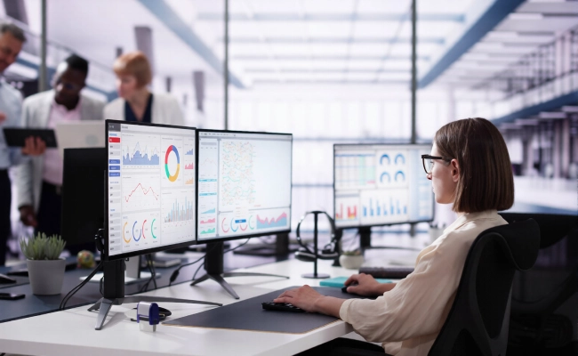Woman in glasses sits at a desk with multiple monitors displaying charts and dashboards in a modern open office environment.