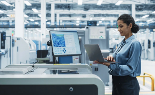 Woman in a blue shirt uses a laptop beside a large manufacturing machine with a monitor in a bright factory setting.